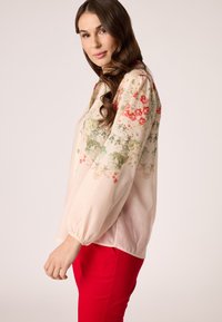 Bloemige blouse van lichtroze stof met pofmouwen. Heeft rode en groene bloemen, een ronde halslijn en een zachte textuur. Gecombineerd met rode broeken.