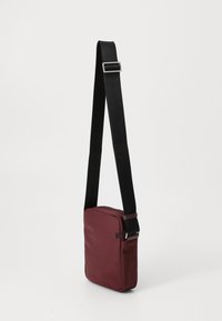 Burgundy crossover taske med en glat overflade, rektangulær form, justerbar sort strop og sølvfarvede hardware detaljer.