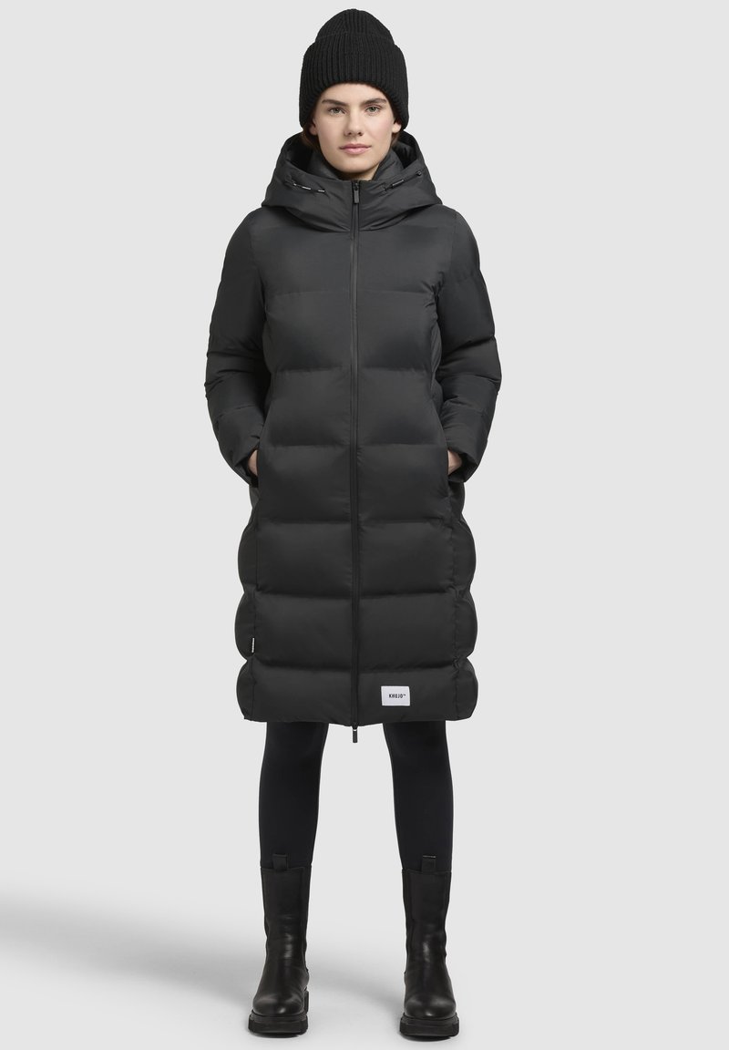 Piumino nero con zip frontale, cappuccio e tasche laterali, caratterizzato da un design trapuntato e una silhouette allungata. Indossato con un berretto nero.