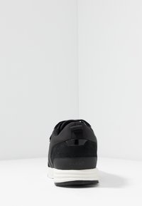 Svart sneaker med vit sula och dragögla sedd från hälen på en vit yta och bakgrund, varumärket "HUGO" synligt.