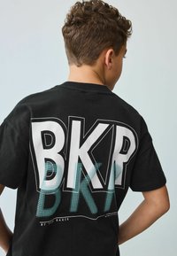 Čierne bavlnené tričko s veľkým bielym a akvamarínovým grafickým dizajnom "BKP" na chrbte, s bodkovými akcentmi a uvoľneným strihom.