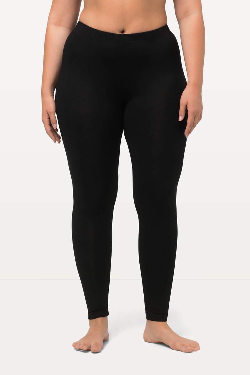Frau trägt hoch taillierte schwarze Leggings, barfuß stehend vor einem einfarbigen weißen Hintergrund.