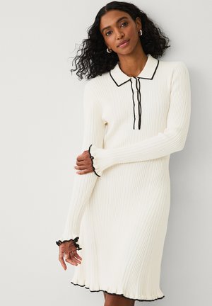 Ribbed white dress featuring a black collar and button placket, with ruffled black accents at the cuffs and hem. Long sleeves.