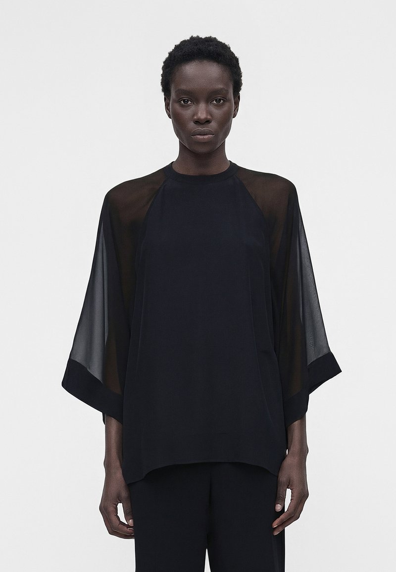 Άτομο που φοράει φαρδύ μαύρο τοπ με διαφανή μακριά μανίκια και φαρδιές μανσέτες, στέκεται μπροστά σε απλό φωτεινό φόντο.