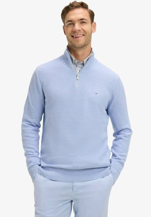 Homme souriant portant un pull à demi-fermeture éclair bleu clair par-dessus une chemise à col rayée, les mains dans les poches, sur un fond blanc.