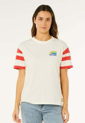 Joven mujer que lleva una camiseta blanca de Rip Curl con rayas rojas en las mangas y vaqueros azules, posando frente a un fondo liso.