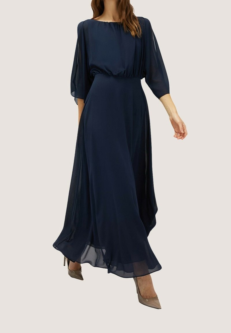 Marineblå maxiklänning gjord av lätt, genomskinligt tyg med en rynkad midja och flödande, vida ärmar. Kompletteras av beige klackar.