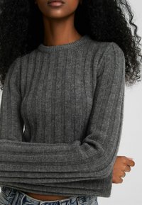 Woman with dark curly hair wearing a fitted grey ribbed sweater and blue jeans, arms crossed in front, cropped below eyes.