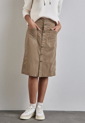 Femme portant une jupe beige boutonnée devant avec de grandes poches avant, des baskets blanches, des chaussettes blanches et un haut à manches longues blanc, debout à l'intérieur.