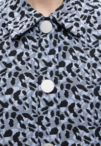 Ανοιχτό μπλε ύφασμα με ακανόνιστα μαύρα λεοπάρ στίγματα, με γιακά και κάθετη σειρά από μεγάλα λευκά κουμπιά.