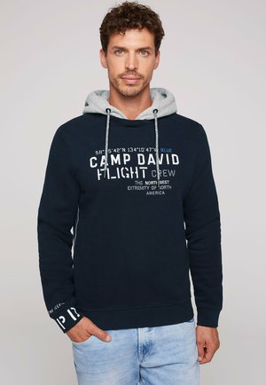 Uomo dai capelli ricci che indossa una felpa con cappuccio blu navy con cappuccio grigio e testo bianco, in piedi con una mano nella tasca dei jeans azzurri.