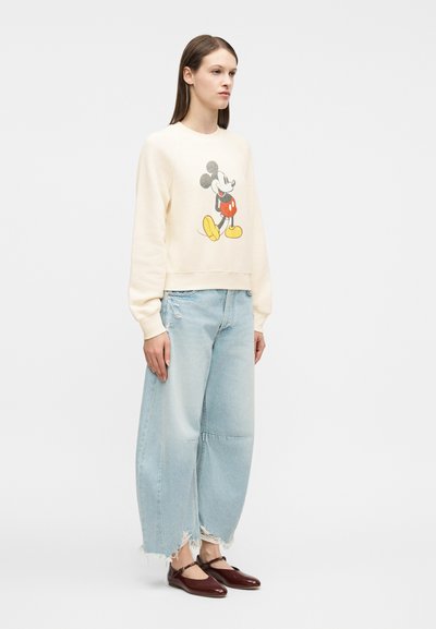 Γυναίκα που φοράει κρεμ φούτερ με εκτύπωση Mickey Mouse, ανοιχτό μπλε φαρδιά φθαρμένα τζιν και σκούρα κόκκινα παπούτσια Mary Jane, στέκεται πλευρικά.