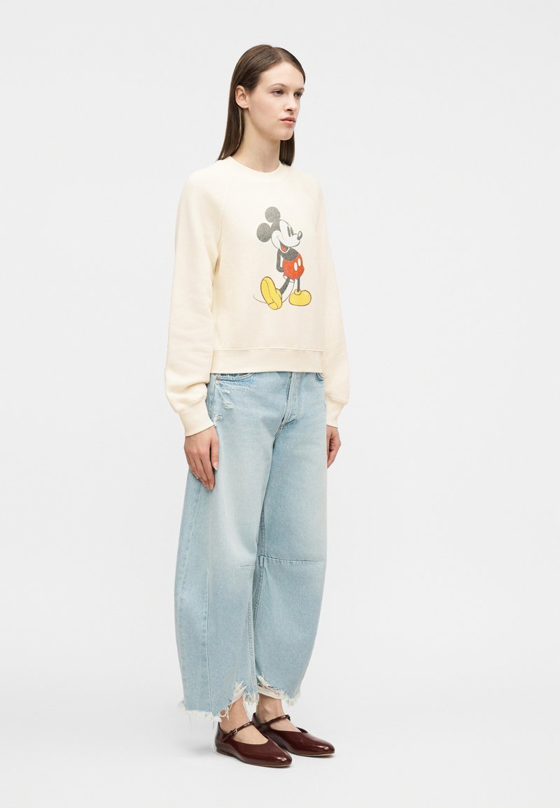 Femme portant un sweat crème avec un motif Mickey Mouse, un jean large déchiré bleu clair et des chaussures Mary Jane bordeaux, debout de profil.