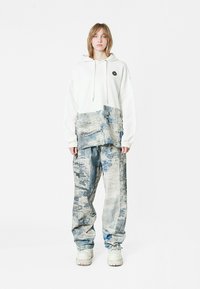 Sudadera blanca de gran tamaño con bolsillo delantero y logo negro, combinada con pantalones baggy azules con estampado. El calzado es de color claro y de suela gruesa.