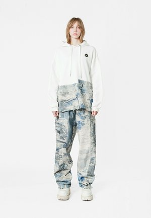 Sudadera blanca de gran tamaño con bolsillo delantero y logo negro, combinada con pantalones baggy azules con estampado. El calzado es de color claro y de suela gruesa.