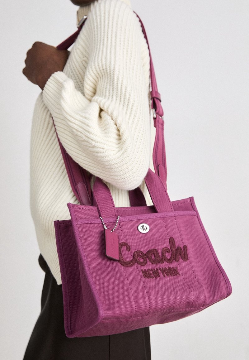 Personne portant un pull crème tient un sac cabas violet Coach avec bandoulière et logo brodé.