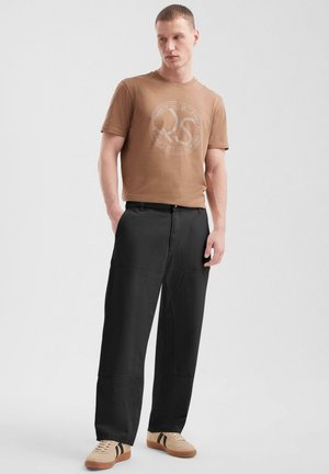 Bruine katoenen t-shirt met een rond logo, gecombineerd met losse zwarte broek en beige sneakers met zwarte strepen. Eenvoudig, casual ontwerp.