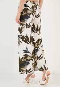 Wijdlopende broek van witte stof met een groen en blauw bladerenpatroon. Elastische tailleband, textuur, gecombineerd met witte hoge hakken.