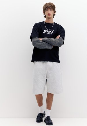 Młody mężczyzna stoi ze skrzyżowanymi rękami, ubrany w czarną koszulkę z nadrukiem na długich szarych rękawach, jasnoszare szorty, białe skarpetki i czarne buty.