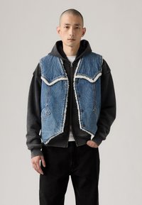 Denimvest med ribbede kanter, lys blå farve, knaplukning foran og spidse yoke-design, lagd over en mørkegrå hættetrøje og sorte bukser.