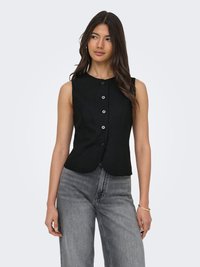 Femme aux longs cheveux foncés portant un gilet noir sans manches boutonné et un jean gris taille haute, debout les mains détendues le long du corps.