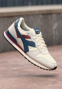 Beige athletic shoe with blue and burgundy accents. Features perforated texture, round toe, and a rubber sole with a textured tread pattern.