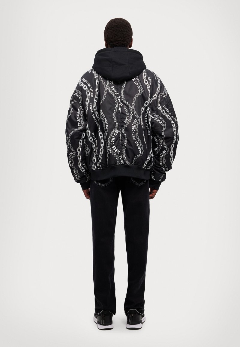 Giacca a vento nera con stampa a motivo catena bianca, dotata di cappuccio, polsini e orlo a costine, abbinata a pantaloni neri e sneakers.