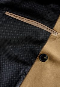 Béžová látka s hladkým povrchom a čiernym hexagonálnym vzorom. Obsahuje zipsovú kapsu a čierny gombík ako akcent.