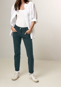 Chino kalhoty z tmavě tyrkysové tkaniny s úzkým střihem. Stylizováno s bílým tílkem a košilí na knoflíky, doplněno bíl�ými teniskami.