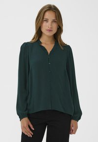 Donkergroene blouse met een gerimpelde halslijn, knoopsluiting aan de voorzijde en lange mouwen, gemaakt van een gestructureerde stof, gedragen met zwarte broek.