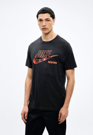 Jeune homme aux cheveux courts et bouclés portant un t-shirt Nike noir avec un logo orange et le texte "Pays-Bas", debout devant un fond blanc.