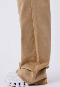 Pantalones de pana marrón claro con una sutil textura acanalada, combinados con zapatillas deportivas blancas que presentan un diseño de suela ondulada único.