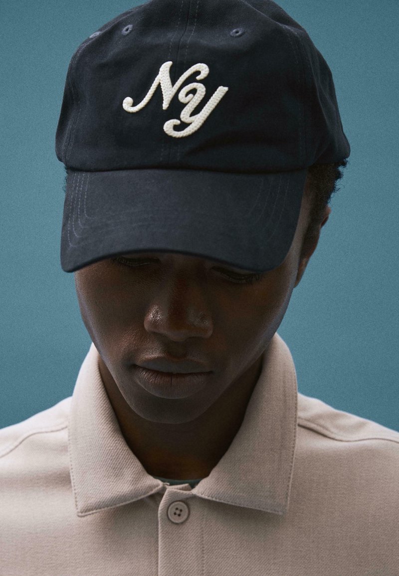 Personne portant une casquette de baseball noire avec le texte blanc "Nóy", chemise beige à col, regardant vers le bas sur un fond bleu uni.