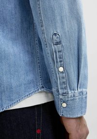 Camicia in denim azzurro chiaro con polsini bottonati con bottoni a forma di perla, orlo curvo e cuciture cucite per un design strutturato.