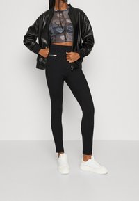 Zwarte faux leren bomberjack over een transparante, geprinte crop top en high-waisted zwarte leggings; witte sneakers maken de outfit compleet.