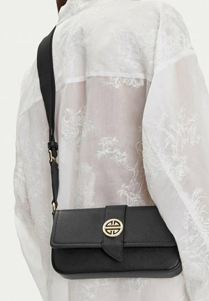 Borsa a tracolla nera con chiusura circolare dorata indossata sulla spalla sopra una camicia bianca trasparente con ricami floreali.