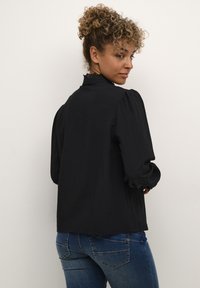 Black blouse with puffed long sleeves, high neckline with lace trim, textured fabric, paired with blue denim jeans. Back view.
