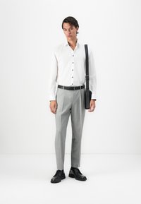 Man wearing white button-up shirt, grey trousers, black belt, black shoes, and carrying a black shoulder bag against a plain white background.