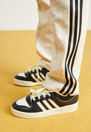 Persoon met zwarte en witte Adidas sneakers en crèmekleurige broek met zwarte strepen, staand op een mosterdgele vloer.