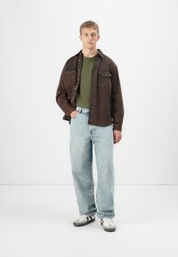 Ung mann iført mørkebrunt overskjorte, grønn T-skjorte, lyseblå jeans og hvite sneakers som står mot en enkel hvit bakgrunn.