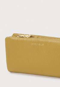 Portofel din piele texturată în nuanță de galben muștar, cu fermoar aurie și numele mărcii Coccinelle embosat în aur pe față.