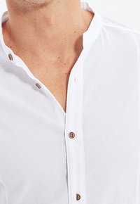 Biała koszula na guziki z tradycyjnym kołnierzykiem i fakturowaną tkaniną. Posiada brązowe guziki na przodzie i przy kołnierzyku.