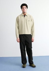 Jachetă crem cu dungi, cu fermoar, dotată cu un buzunar pe piept, și pantaloni negri. Modelul poartă pantofi verzi din material camuflaj. Fundal simplu.