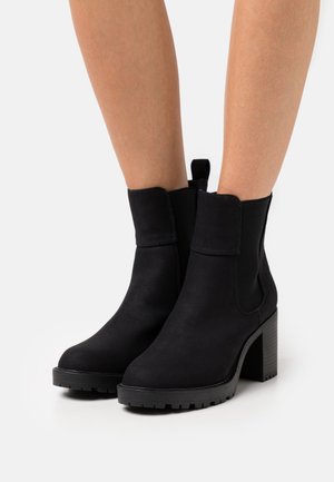 Sorte suede ankelstøvler med blokhæle og elastiske sidestykker, båret på bare ben mod en hvid baggrund.