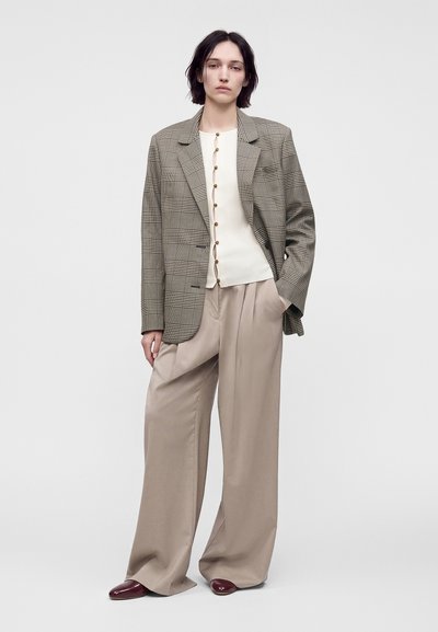 Femme portant un pantalon beige large, un haut blanc boutonné, un blazer à carreaux et des chaussures rouge foncé, debout devant un fond clair uni.