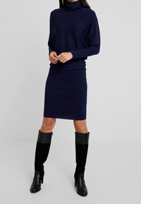 Laivastonsininen poolopaita, jossa on kiinteä malli, yhdistettynä matching polvenpituiseen hameeseen. Mallilla on päällä mustat polvisukat, joissa on sileä viimeistely.