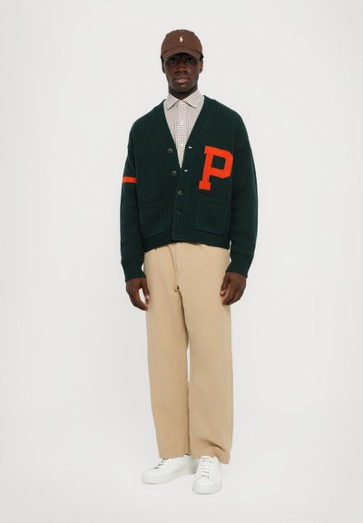Cárdigan de punto verde con la letra "P" naranja y raya, botones negros, llevado sobre una camisa de cuadros, combinado con pantalones beiges y zapatillas blancas.