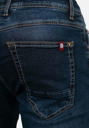 Donker denim jeans met een achterzak met oranje stiksel en een klein rood-wit merklabel. De stof heeft een gladde textuur.