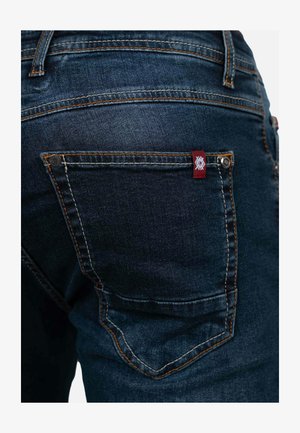 Donker denim jeans met een achterzak met oranje stiksel en een klein rood-wit merklabel. De stof heeft een gladde textuur.
