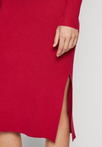 Robe rouge en tricot avec texture côtelée, manches longues et fente latérale. Détail d'une main visible près de l'ourlet de la robe.